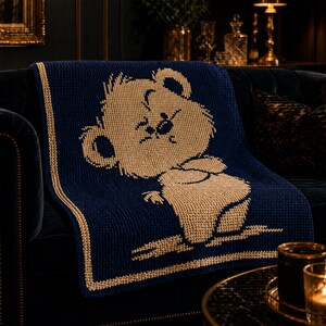 May include: A navy blue knitted throw blanket with a tan cartoon bear design. The blanket has a tan border and is draped over a dark blue sofa. The bear is in a seated position with its arms crossed.