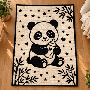 May include: A rectangular crochet blanket featuring a cream-colored background with a black border. The center displays a black and white panda holding a baby bottle, surrounded by black stars, hearts, and bamboo leaf patterns.