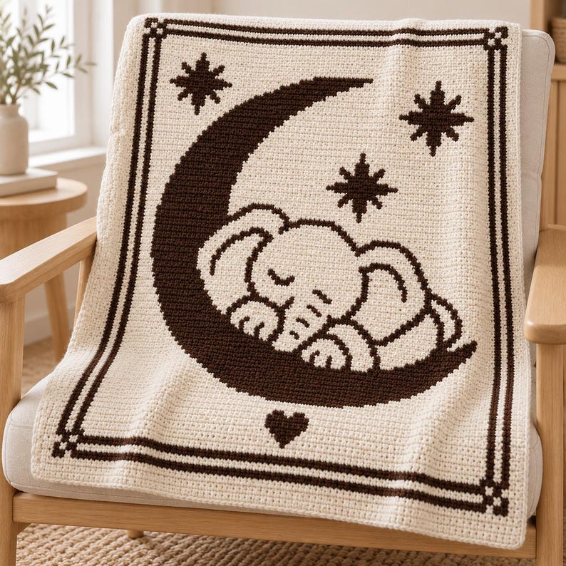 May include: A cream-colored crochet blanket featuring a dark brown border and a central graphic of a sleeping elephant resting on a crescent moon. The design includes three dark brown stars and a small heart shape, all rendered in a textured stitch.