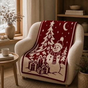 May include: A rectangular knit throw blanket featuring a deep red background with a cream-colored pattern. The design displays a snowman wearing a hat and scarf, a decorated Christmas tree, stacked gift boxes, a crescent moon, and small stars.