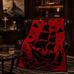 May include: A rectangular knit blanket featuring a bold red background with a large black graphic of a sailing ship on turbulent waves. The design includes black silhouettes of birds and clouds in the sky, finished with a solid black border.