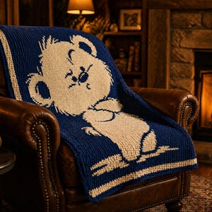 May include: A navy blue and cream knitted blanket draped over a brown leather armchair. The blanket features a large bear design in cream against the navy blue background. The bear is sitting down. The blanket has cream stripes.