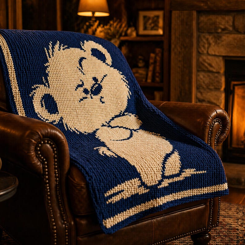 May include: A navy blue and cream knitted blanket draped over a brown leather armchair. The blanket features a large bear design in cream against the navy blue background. The bear is sitting down. The blanket has cream stripes.