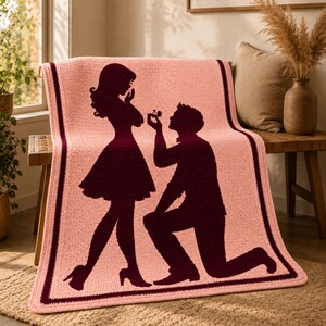 May include: A pink crocheted blanket featuring a silhouette of a proposal scene. The design shows a man kneeling and offering a ring to a woman. The blanket has a dark burgundy border.