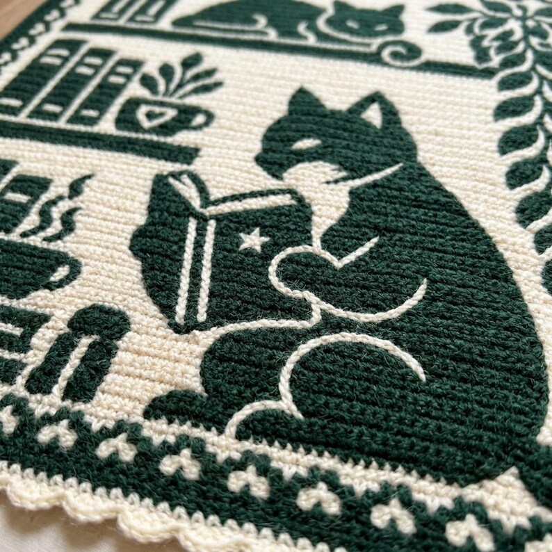 May include: A cream-colored crochet textile featuring a dark green graphic design of a cat reading a book with a star on the cover. The scene includes shelves with books, a potted plant, a steaming mug, and a decorative border pattern.