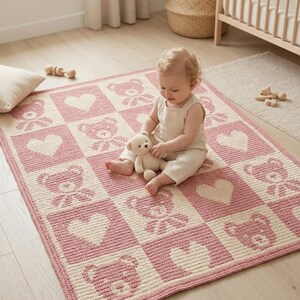 May include: A pink and cream-colored baby blanket with a repeating pattern of teddy bears and hearts. The blanket is made of a soft, knitted material and is rectangular in shape. A baby is sitting on the blanket, holding a teddy bear.