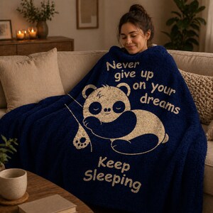 May include: A navy blue sherpa blanket featuring a sleeping panda illustration and the text "Never give up on your dreams" and "Keep Sleeping" in cream. The blanket is draped over a person on a beige sofa.