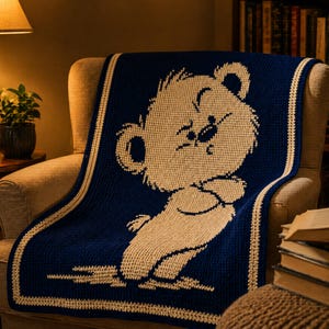 May include: A navy blue and cream crocheted blanket draped over a chair. The blanket features a large, smiling bear design in cream against a navy blue background. The blanket has a cream border.