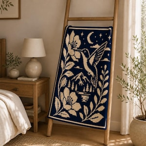 May include: A rectangular knit throw blanket featuring a dark navy blue background with a cream-colored graphic design. The pattern depicts a hummingbird hovering near a large flower, a crescent moon, stars, mountain peaks, and pine trees.