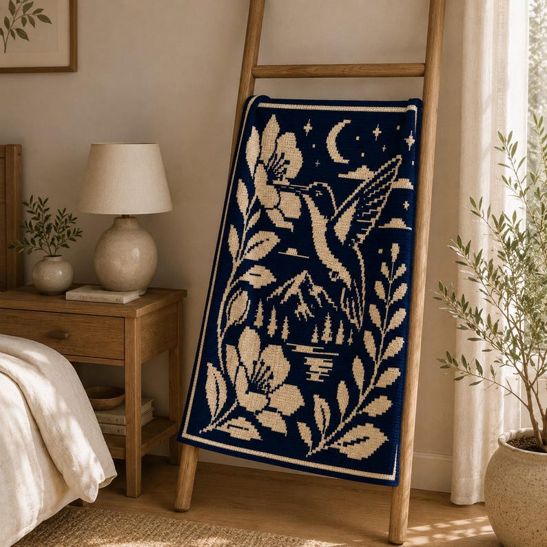 May include: A rectangular knit throw blanket featuring a dark navy blue background with a cream-colored graphic design. The pattern depicts a hummingbird hovering near a large flower, a crescent moon, stars, mountain peaks, and pine trees.