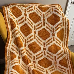 May include: A crochet blanket featuring a repeating geometric pattern of white lines on a mustard yellow background. The design consists of interconnected diamond and square shapes, finished with a simple white and yellow border.