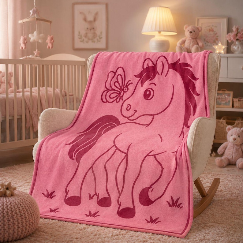 May include: A pink baby blanket with a cartoon horse and butterfly design. The blanket is displayed on a rocking chair in a nursery. The horse illustration is in a darker shade of pink.