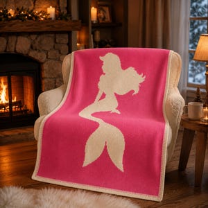 May include: A pink throw blanket with a cream-colored mermaid silhouette design. The blanket is draped over a cream-colored armchair. The blanket has a cream-colored border.