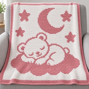 May include: A white and pink crocheted blanket featuring a central graphic of a sleeping bear resting on a cloud. The design includes a crescent moon and four stars in pink, with a solid pink border framing the entire rectangular piece.
