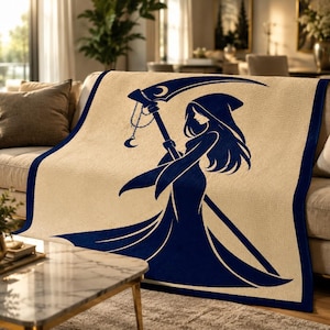 May include: A rectangular throw blanket featuring a cream-coloured base with a dark navy blue border and a large, central graphic illustration of a hooded figure in a flowing gown holding a scythe with a crescent moon charm hanging from the handle.