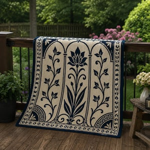 May include: A beige and navy blue outdoor rug with a floral design draped over a wooden deck railing. The rug features a central lotus flower motif and scrolling leaf patterns within arched panels. The border is navy blue with geometric details.
