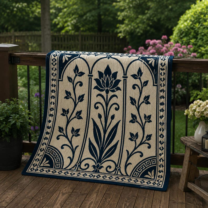 May include: A beige and navy blue outdoor rug with a floral design draped over a wooden deck railing. The rug features a central lotus flower motif and scrolling leaf patterns within arched panels. The border is navy blue with geometric details.