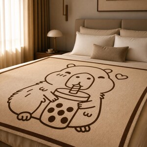 May include: A beige blanket with a brown border featuring a cartoon hamster holding a boba tea. The hamster is outlined in brown and has a cute expression. The blanket is on a bed with white pillows and a brown pillow.