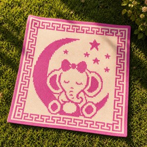 May include: A square baby blanket with a pink and white elephant design. The elephant is sitting on a crescent moon with stars. The blanket has a pink border with a Greek key pattern. The blanket is on a green surface.