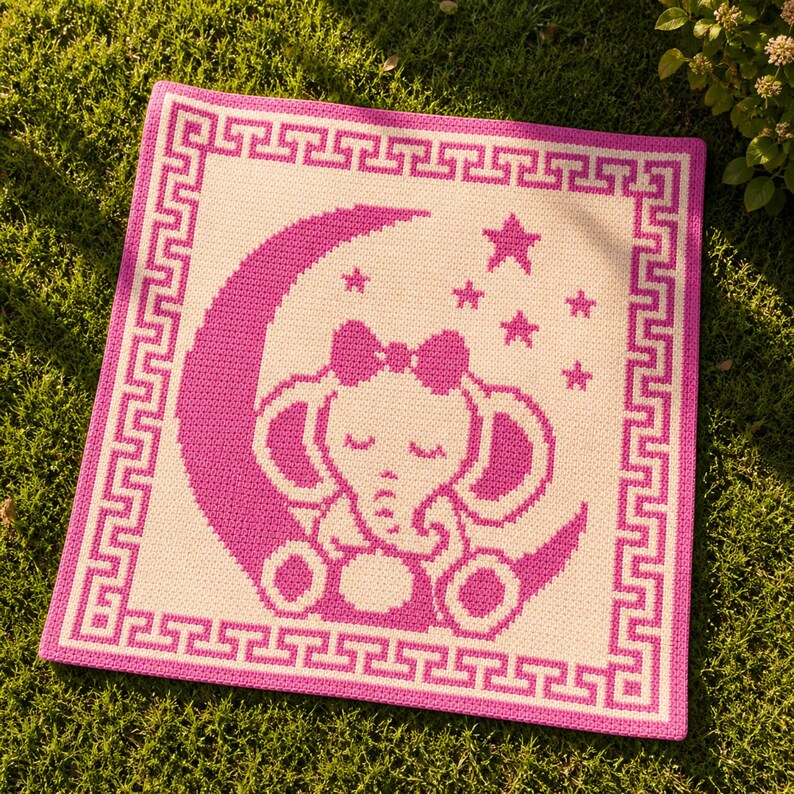 May include: A square baby blanket with a pink and white elephant design. The elephant is sitting on a crescent moon with stars. The blanket has a pink border with a Greek key pattern. The blanket is on a green surface.