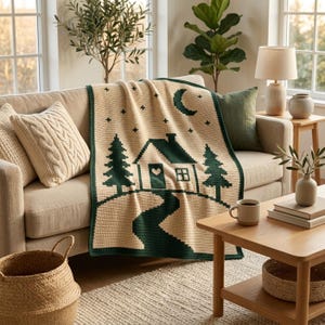Pode incluir: Uma manta de croch&ecirc; na cor creme com um design gr&aacute;fico em verde escuro que apresenta uma cabana, pinheiros, uma lua crescente e estrelas. A cabana possui um detalhe em formato de cora&ccedil;&atilde;o na porta e uma janela.