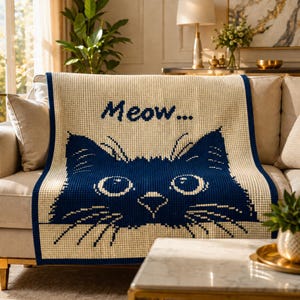 Könnte beinhalten: Eine cremefarbene Decke mit einem marineblauen Katzen-Gesicht-Design und dem Wort "Meow...". Die Decke ist über ein beiges Sofa drapiert. Die Augen und die Nase der Katze sind weiß, und die Schnurrhaare sind marineblau.