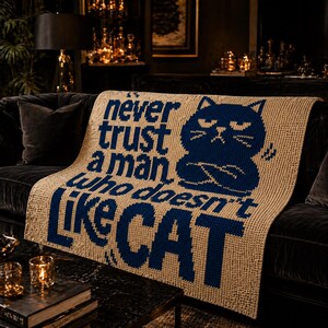 May include: Beige knitted throw blanket with the text "Never trust a man who doesn't like cat" in navy blue. A navy blue cat graphic is also featured on the blanket. The blanket is draped over a dark sofa.