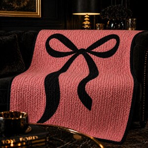 Pu&ograve; includere: Una coperta lavorata a maglia in una tonalit&agrave; rosa tenue, con un grande disegno a fiocco nero. La coperta &egrave; drappeggiata su un divano scuro, mostrando la sua maglia strutturata e i colori contrastanti. Questo plaid decorativo aggiunge un tocco di eleganza a qualsiasi spazio abitativo.