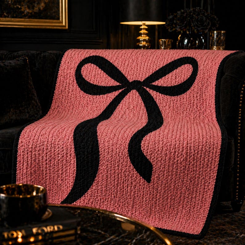 Pu&ograve; includere: Una coperta lavorata a maglia in una tonalit&agrave; rosa tenue, con un grande disegno a fiocco nero. La coperta &egrave; drappeggiata su un divano scuro, mostrando la sua maglia strutturata e i colori contrastanti. Questo plaid decorativo aggiunge un tocco di eleganza a qualsiasi spazio abitativo.