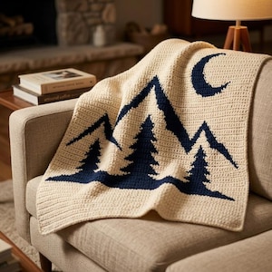 May include: A cream-coloured crochet blanket featuring a dark navy blue graphic design of a mountain range, three pine trees, and a crescent moon. The textile has a visible stitched texture and is draped over a neutral-toned upholstered furniture piece.