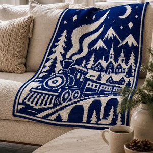May include: A rectangular knit throw blanket featuring a two-tone blue and white pattern. The design depicts a steam train traveling across a bridge toward a village with mountains, stars, and a crescent moon in the night sky above.