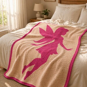 May include: A crocheted blanket featuring a pink fairy silhouette on a beige background. The blanket has a pink border and is displayed on a bed with white bedding. The fairy has outstretched arms and wings.
