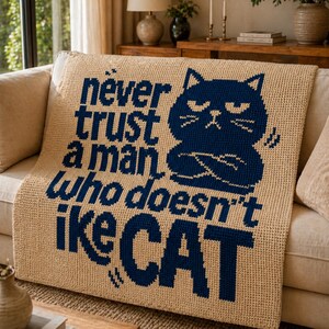 May include: Beige crocheted throw blanket with navy blue text and a cat graphic. The text reads "never trust a man who doesn't like CAT". The cat graphic has crossed arms and a grumpy expression. The blanket is draped over a beige sofa.