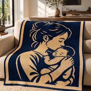 May include: A navy blue and tan throw blanket featuring an illustration of a mother kissing a baby. The blanket is draped over a beige sofa. The illustration is in tan, with a navy blue background and border.