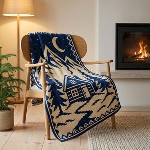 May include: A navy blue and cream knit throw blanket featuring a repeating pattern of a cabin, pine trees, mountains, and a crescent moon. The blanket is draped over a light-colored wooden chair, showcasing a detailed winter-themed graphic design.