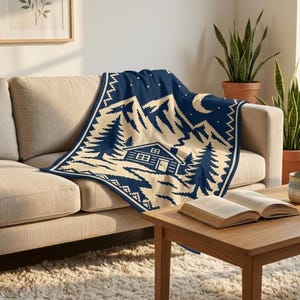 May include: A navy blue and cream-colored knit throw blanket featuring a repeating pattern of a cabin, pine trees, mountains, and a crescent moon. The blanket is draped over a beige sofa, showcasing a detailed landscape design with a border.