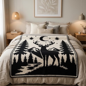 Peut inclure: Un plaid rectangulaire pr&eacute;sentant un motif tricot&eacute; contrast&eacute; en noir et cr&egrave;me. Le design repr&eacute;sente la silhouette centrale d'un cerf devant une cha&icirc;ne de montagnes, entour&eacute; de sapins, d'un croissant de lune et de plusieurs &eacute;toiles.