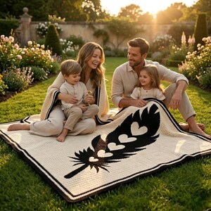 May include: A family is seated on a cream blanket featuring a black feather design with heart motifs. The blanket has a black and brown border. They are on a lawn, surrounded by flowers and trees in a garden setting.