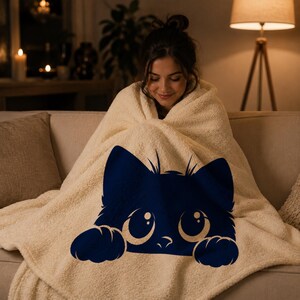 May include: A cream-colored sherpa blanket featuring a large navy blue cat face design. The cat has big, round eyes and paws. The blanket is draped over a person sitting on a beige couch in a cozy, dimly lit room.