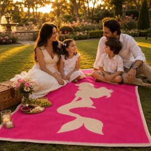 May include: A bright pink blanket with a white mermaid silhouette is spread on the grass. A family is seated around the blanket. A picnic basket, flowers, and snacks are nearby. The blanket has a white border.