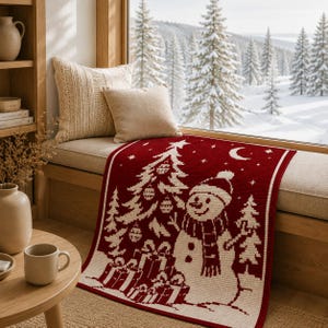 May include: A rectangular knit throw blanket featuring a deep red background with a cream-colored pattern of a snowman, a decorated Christmas tree, wrapped gifts, stars, and a crescent moon, displayed on a window seat with two beige pillows.