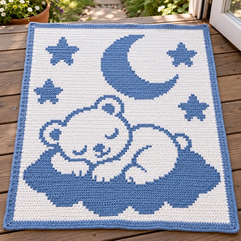 May include: A rectangular crochet blanket featuring a white background with a blue border. The design displays a white sleeping bear resting on a blue cloud, surrounded by a large blue crescent moon and three blue stars in a pixelated style.
