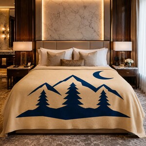 May include: A tan-coloured bed blanket featuring a dark blue graphic print of a mountain range, three pine trees, and a crescent moon. The blanket is draped over a bed with white pillows and a headboard in a bedroom setting with two bedside lamps.