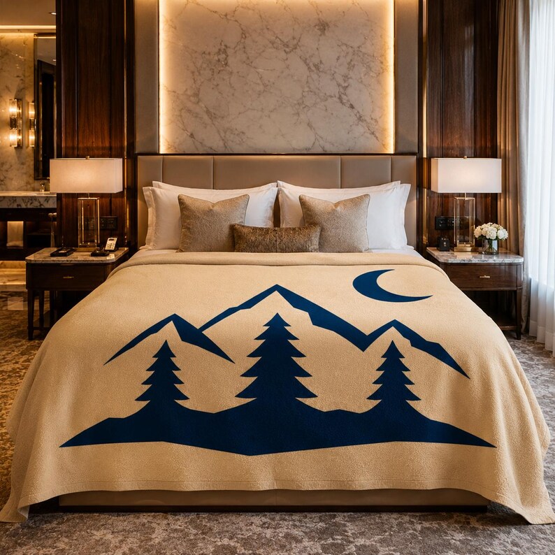 May include: A tan-coloured bed blanket featuring a dark blue graphic print of a mountain range, three pine trees, and a crescent moon. The blanket is draped over a bed with white pillows and a headboard in a bedroom setting with two bedside lamps.