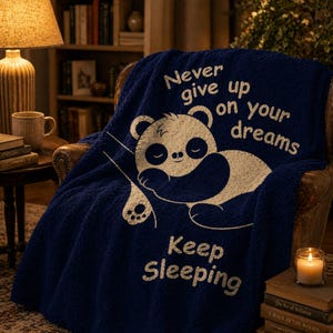 May include: A navy blue sherpa blanket featuring a sleeping panda illustration and the text "Never give up on your dreams, Keep Sleeping." The blanket is displayed on a brown leather armchair, creating a cosy and inviting atmosphere.