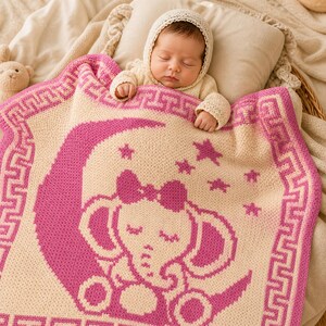 May include: A sleeping baby is nestled under a cream and pink knitted blanket. The blanket features a pink elephant design with a bow, moon, and stars. The blanket has a Greek key border.