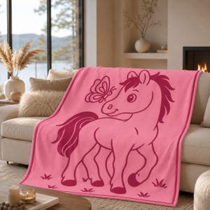May include: A pink throw blanket with a cartoon horse and butterfly design. The horse is a darker shade of pink. The blanket is draped over a beige sofa. The blanket has a border and is decorated with grass.