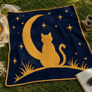 May include: A navy blue crocheted blanket featuring a golden cat silhouette seated on a crescent moon, surrounded by stars. The blanket has a golden border and is placed on green grass. A cozy, handmade item.