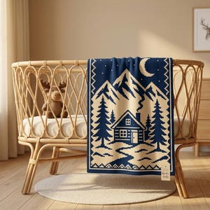 May include: A navy blue and cream knit blanket featuring a mountain landscape, a cabin, pine trees, and a crescent moon. The blanket is draped over a light brown rattan crib. A small tag with the text Nordic Knits is attached to the bottom corner.