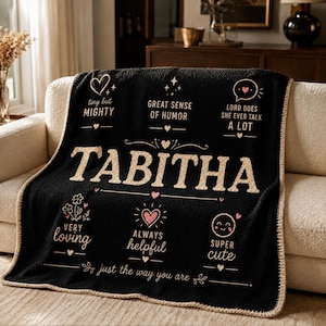 Puede incluir: Manta negra con el nombre "Tabitha" en letras beige. La manta está decorada con frases como "tiny but mighty" y "always helpful", junto con iconos de corazón y estrella.